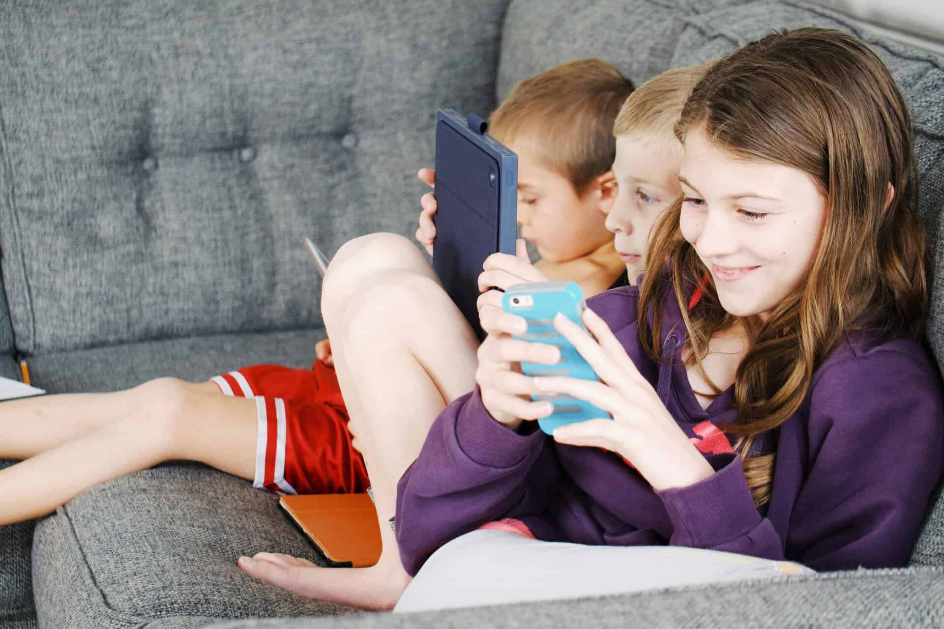 Three children sitting on a couch, engaged with electronic devices, smiling and enjoying leisure time, devices and screen time.