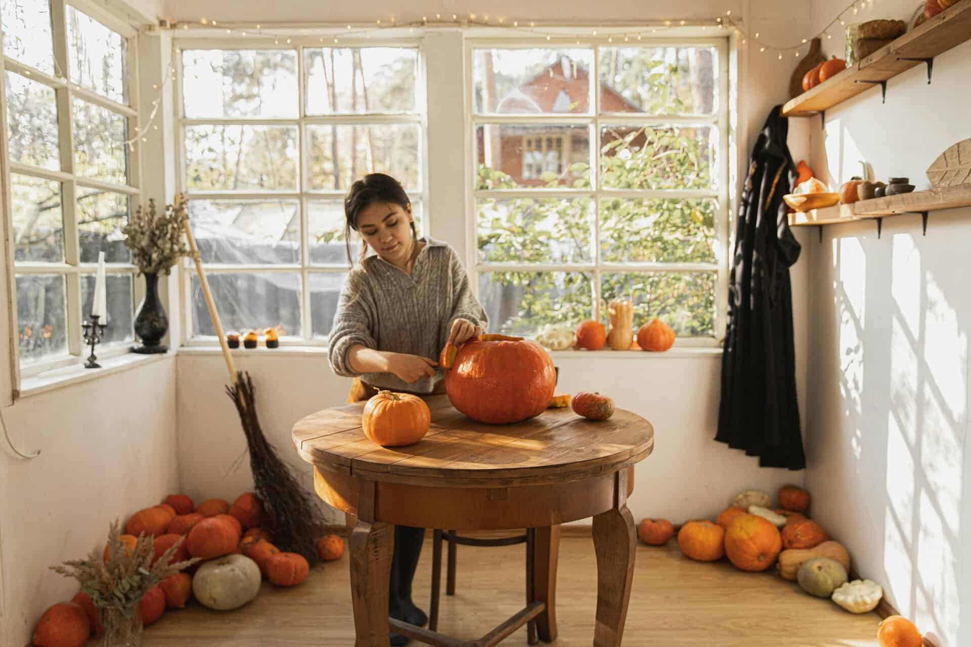 A woman carves pumpkins in a cozy room filled with autumnal decor and natural light.