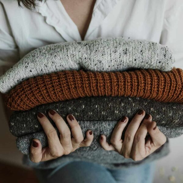 A person holding a stack of folded knitted sweaters in various colors, with a focus on textiles and textures. Fall fashion, fashion trend