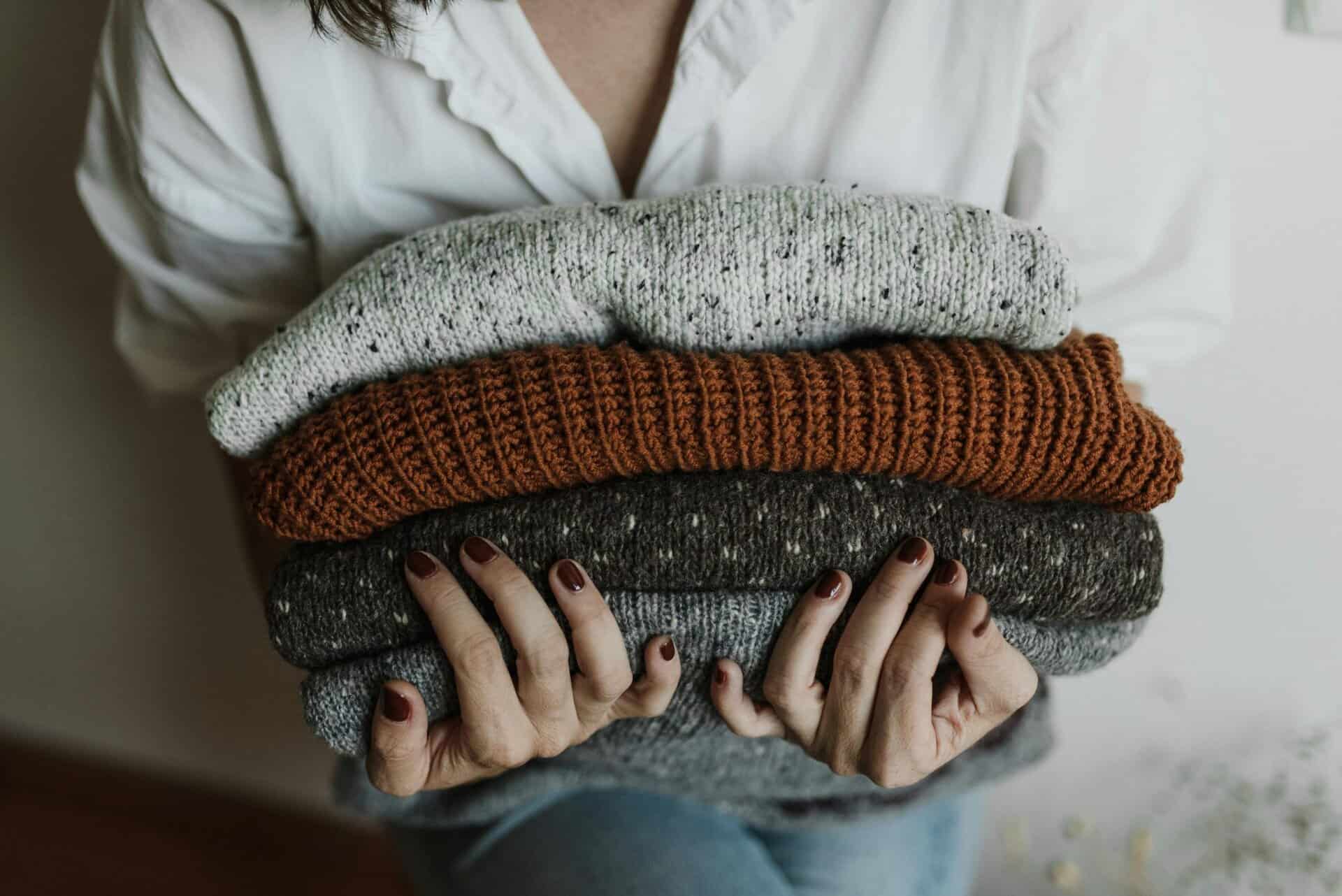 A person holding a stack of folded knitted sweaters in various colors, with a focus on textiles and textures. Fall fashion, fashion trend