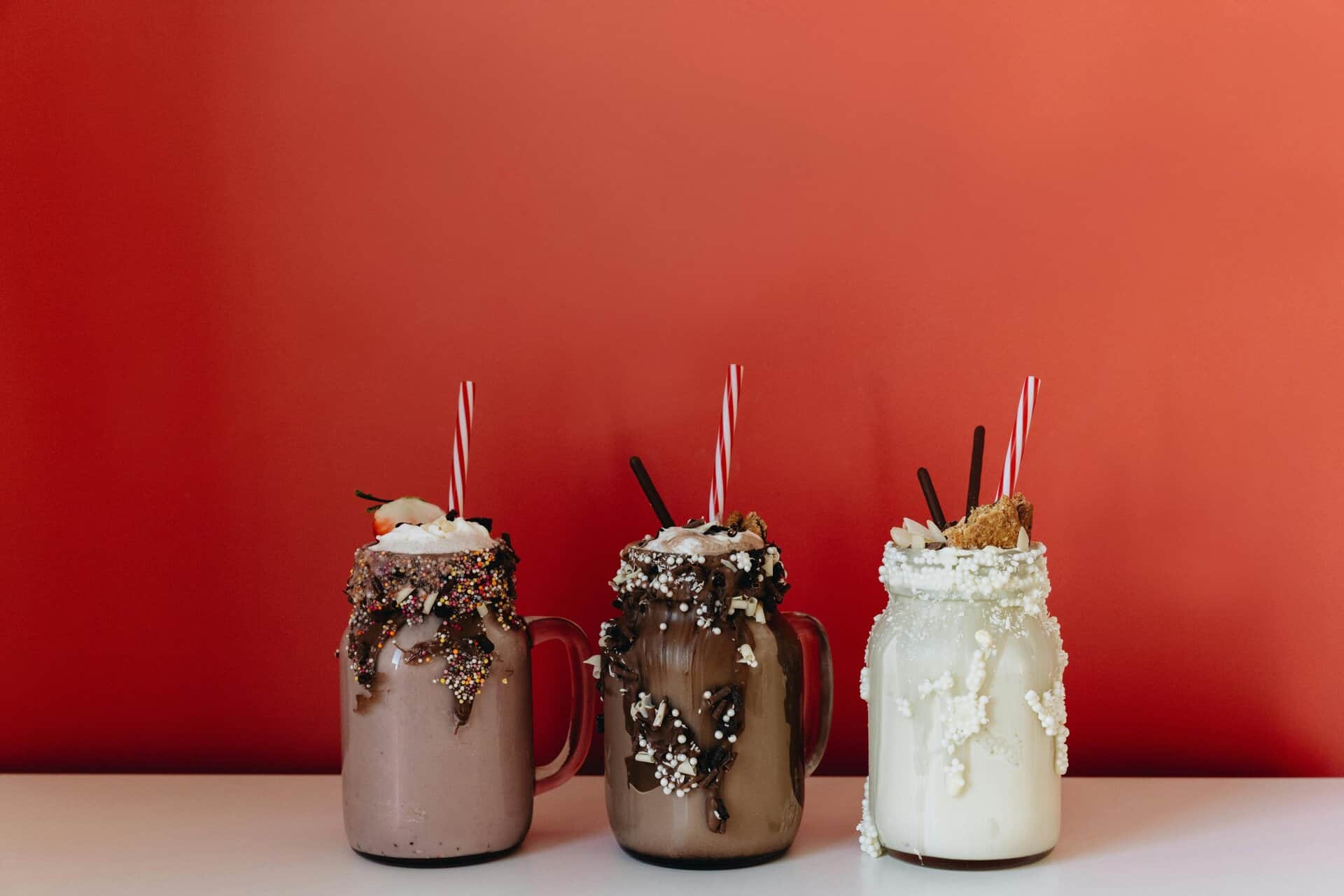 Three indulgent milkshakes in glass jars with sprinkles and chocolate against a bold red backdrop. Viral drink.