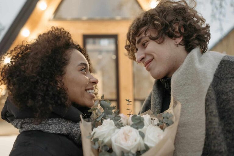 A happy couple smiling at each other with a bouquet of flowers outside a cozy house. Strengthen your relationship and intimacy.