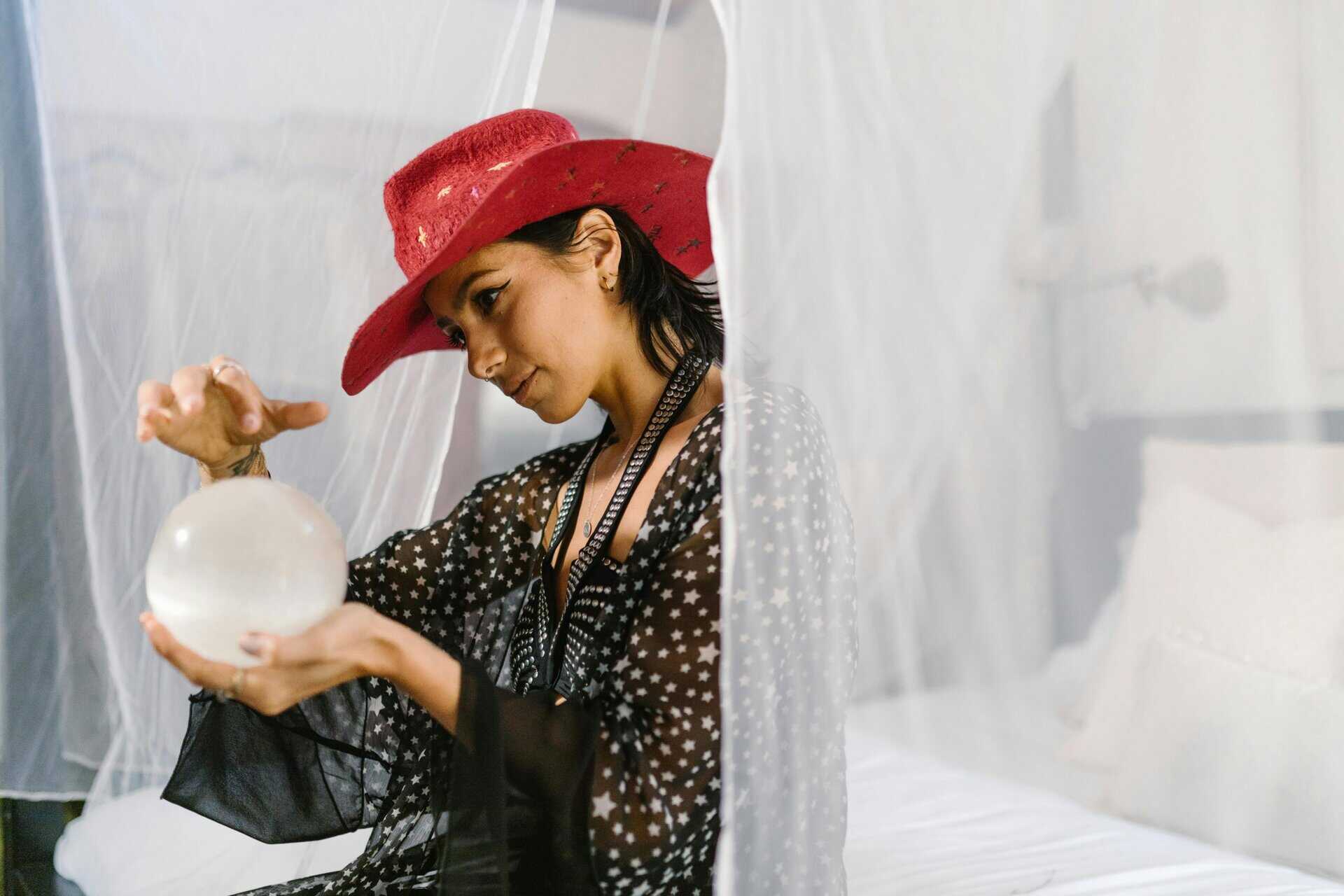 A woman in a red hat examines a crystal ball in a mystical setting, evoking spiritual themes.
