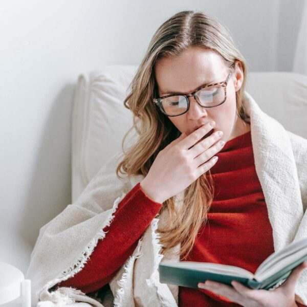 Tired young lady with long blond hair in warm clothes and eyeglasses yawning and covering mouth with hand while reading book sitting in armchair at home
