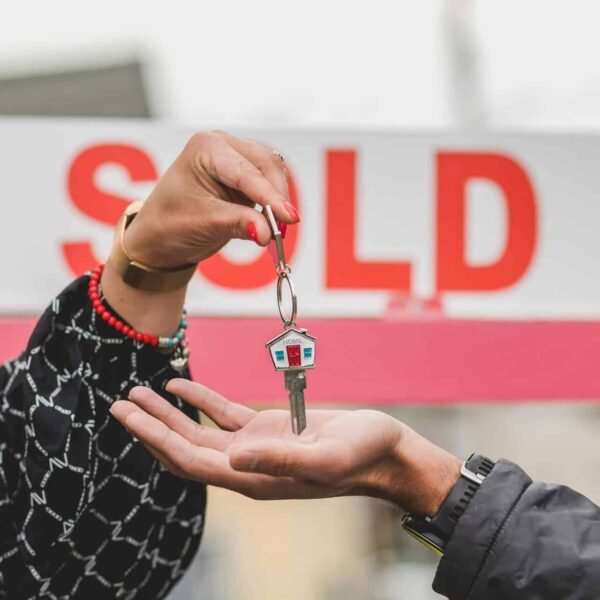 Close-up of handing over house key with sold sign in background, symbolizing real estate success. selling, home value