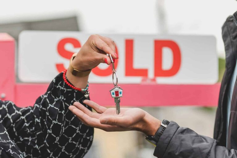 Close-up of handing over house key with sold sign in background, symbolizing real estate success. selling, home value