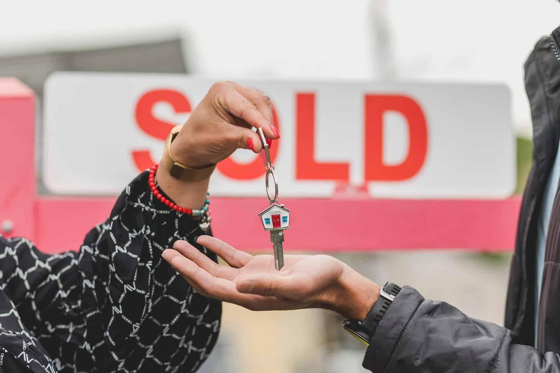 Close-up of handing over house key with sold sign in background, symbolizing real estate success. selling, home value