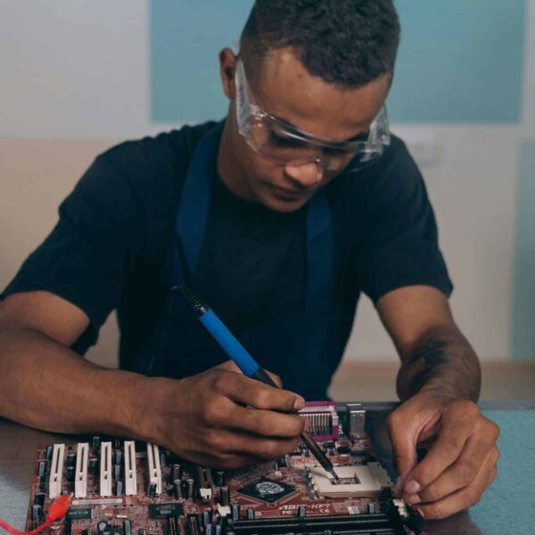 A technician wearing safety glasses works intently on a computer motherboard with a soldering iron. AI Data Centers