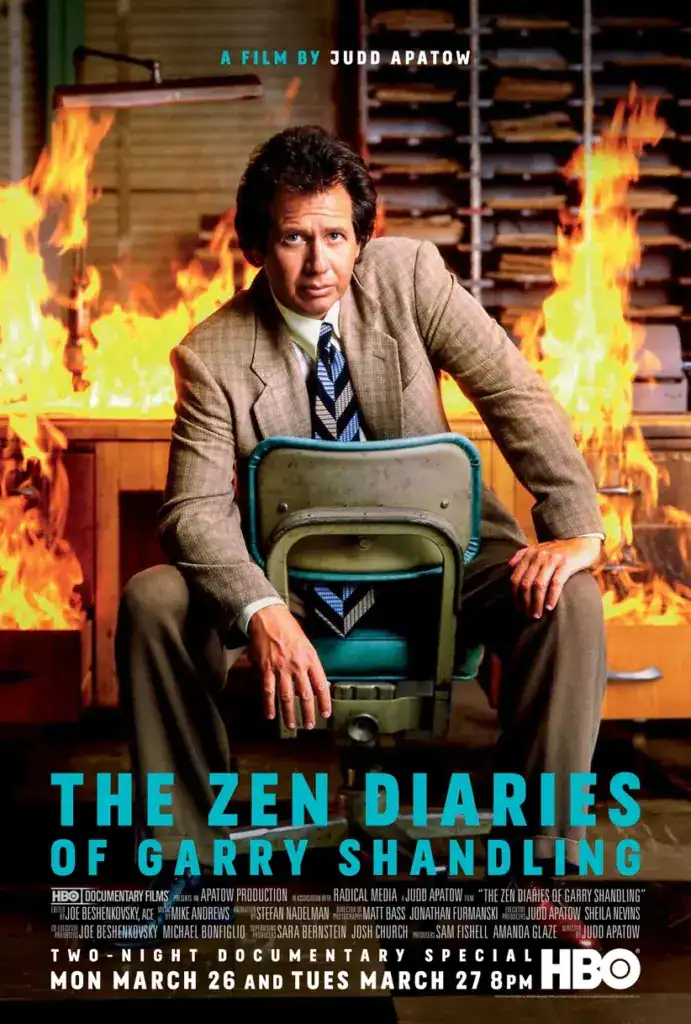 Gary Shandling in a suit, sitting on a chair with a fire behind him.