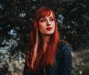 A captivating portrait of a redhead woman standing outdoors among snowflakes, wearing a black jacket. cold weather beauty mistakes to avoid