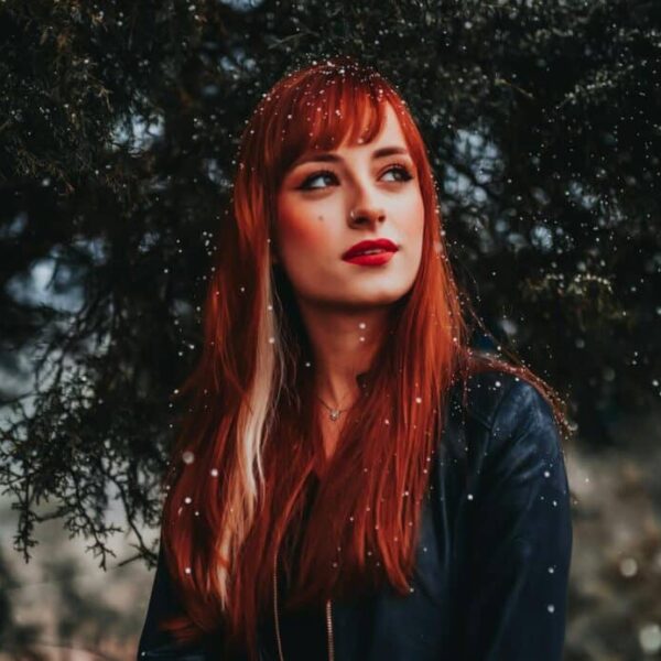 A captivating portrait of a redhead woman standing outdoors among snowflakes, wearing a black jacket. cold weather beauty mistakes to avoid