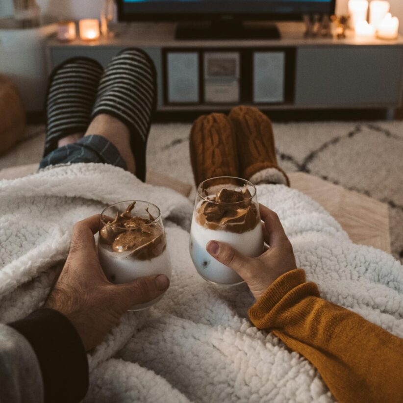 A couple enjoys a cozy movie night with fall mocktails, wrapped in blankets, watching a film on TV.