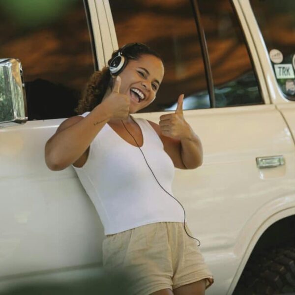 Excited young ethnic female camper showing thumbs up and listening to music in headphones while leaning on car under tent during summer holidays. Hakuna Matata, mental wellness