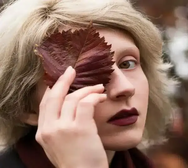 person covering their face with a leaf wearing dark lipstick perfect choice for a good fall lip color. fall beauty