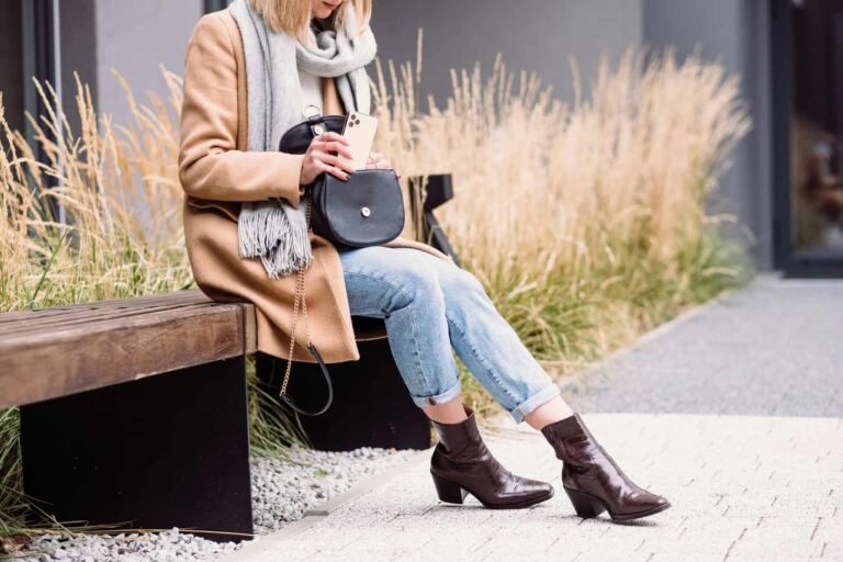 Elegant woman in a coat and scarf using her phone on a bench outdoors in Warsaw. fall boots