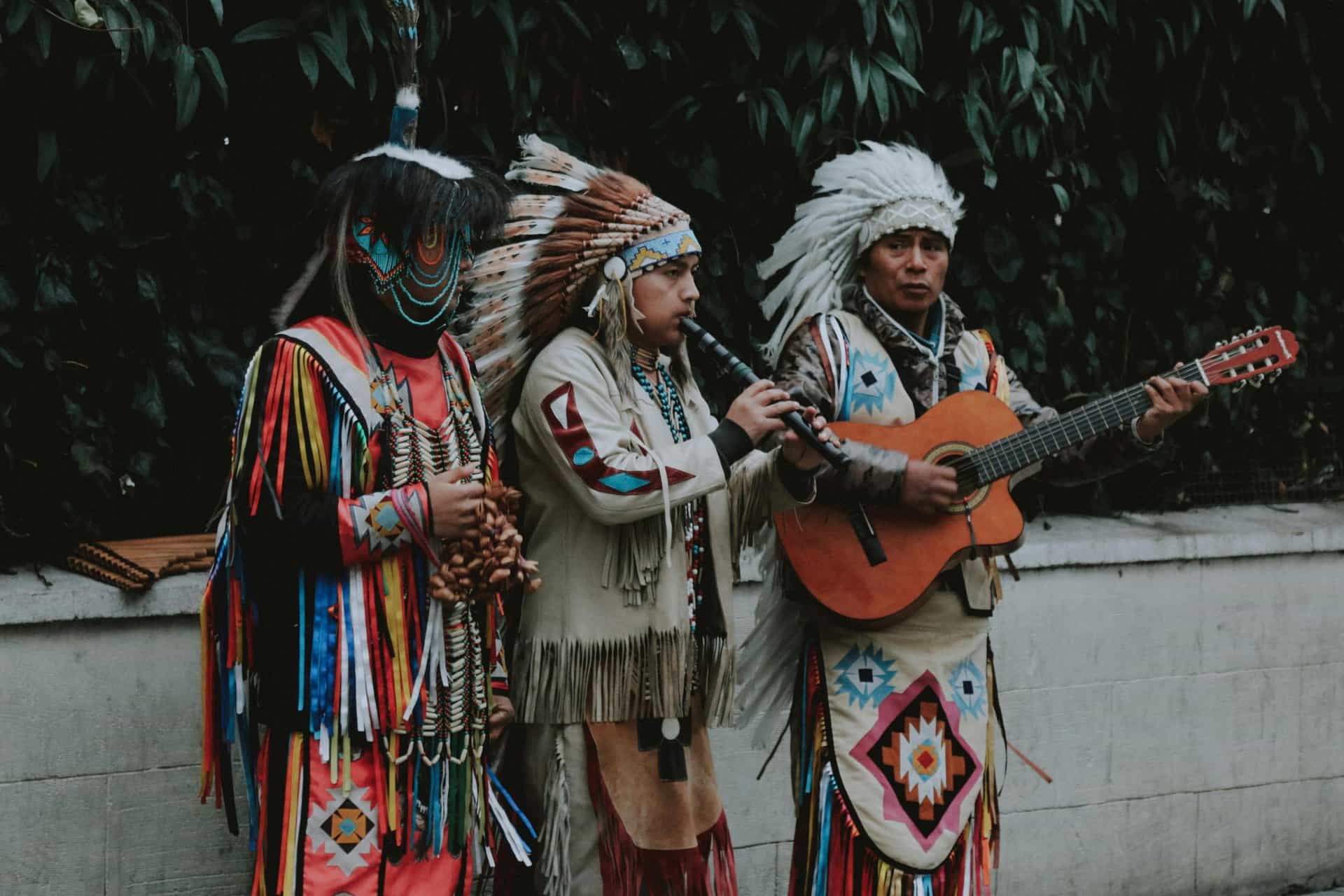 Native American musicians in traditional attire playing flute and guitar outdoors.