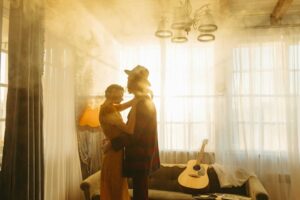 A couple embraces in a warm, sunlit living room with a guitar resting on the sofa. Gratitude, fall songs,