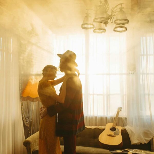 A couple embraces in a warm, sunlit living room with a guitar resting on the sofa. Gratitude, fall songs,