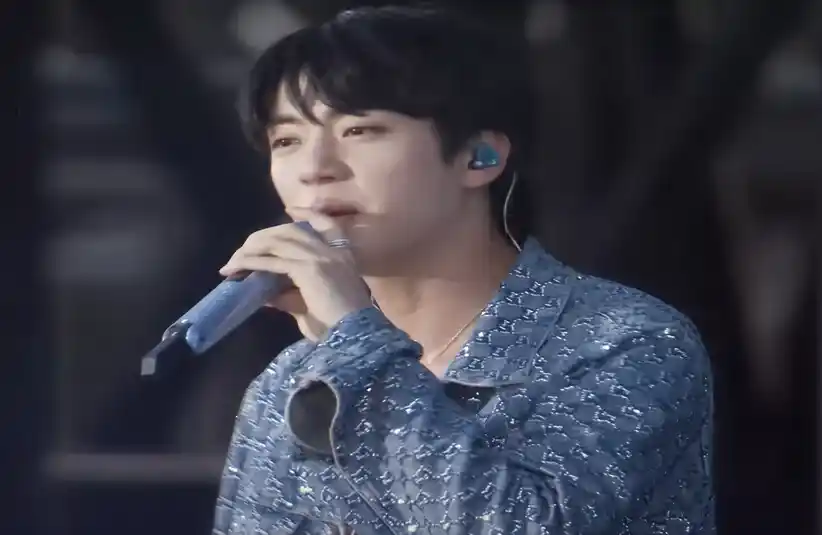 Jin from BTS, sings into a microphone during his first solo tour since his return, while wearing a blue patterned jacket. They have black hair and appear to be performing on stage, conveying a focused expression.