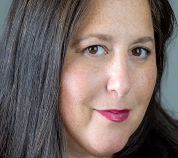 Sundance Comms Head Tammie Rosen, with long dark hair and light makeup, including red lipstick, gazes confidently at the camera. The background is a neutral gray, creating a professional tone.
