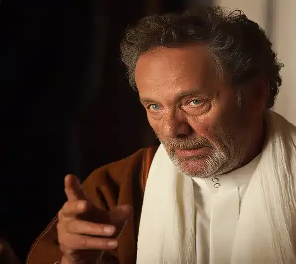 Mohammad Bakri, the filmmaker, playing an older man with curly gray hair gestures thoughtfully. He wears a white robe with a brown cloak. The background is dimly lit, creating an introspective mood.