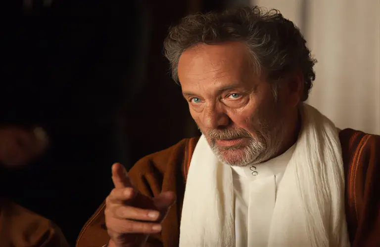 Mohammad Bakri, the filmmaker, playing an older man with curly gray hair gestures thoughtfully. He wears a white robe with a brown cloak. The background is dimly lit, creating an introspective mood.