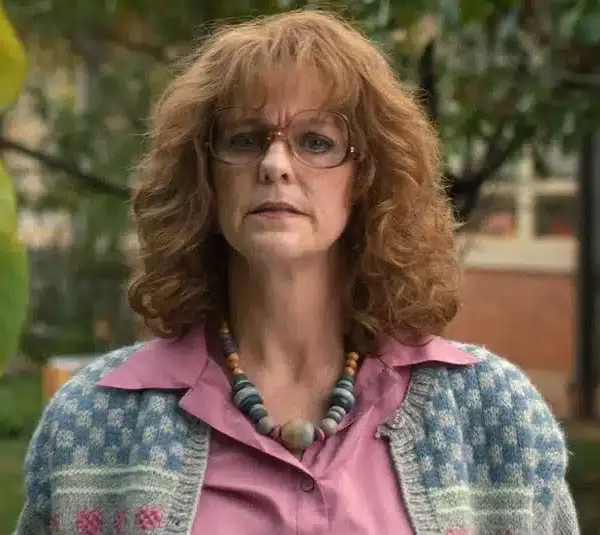 Hope Hynes Love, who plays Miss Harris in Stranger Things, with curly hair and glasses, looks directly at the camera. She's wearing a patterned cardigan, a pink shirt, and a beaded necklace. A tree and a building are in the background.