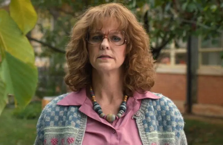 Hope Hynes Love, who plays Miss Harris in Stranger Things, with curly hair and glasses, looks directly at the camera. She's wearing a patterned cardigan, a pink shirt, and a beaded necklace. A tree and a building are in the background.