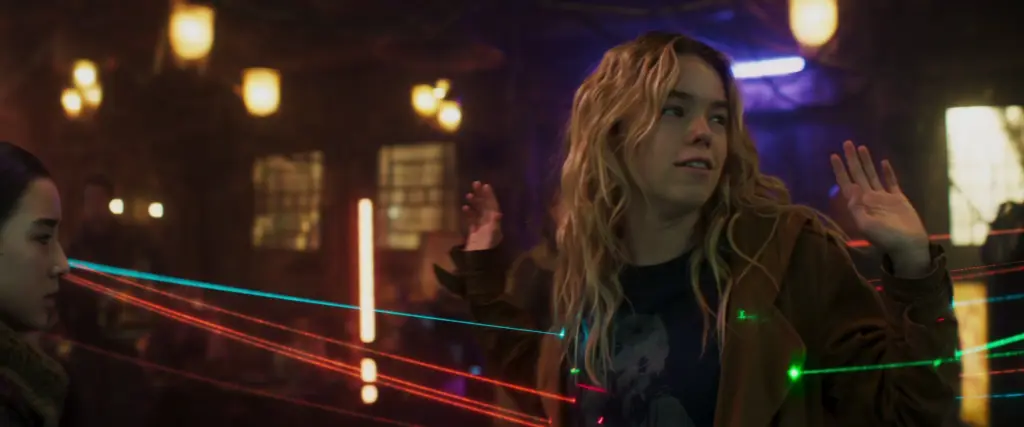 Supergirl with wavy hair stands with hands raised in a dim, neon-lit room, crossed by colorful laser beams, conveying a playful atmosphere.