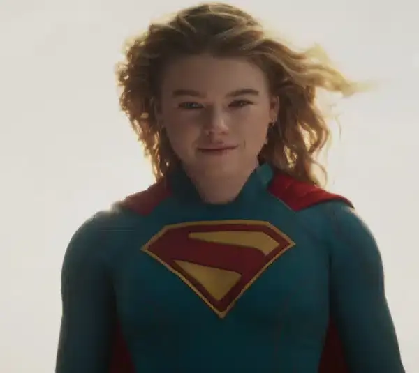 Supergirl played by Milly Alcock, wearing a blue superhero suit with a red and yellow "S" emblem, stands against a bright, cloudy sky. The mood is empowering.