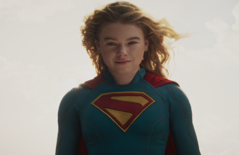 Supergirl played by Milly Alcock, wearing a blue superhero suit with a red and yellow "S" emblem, stands against a bright, cloudy sky. The mood is empowering.
