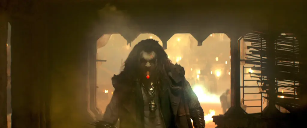 Lobo, played by Jason Momoa, stands in a dimly lit room, exuding a mysterious and intense vibe. Warm lights glow through a window behind them.