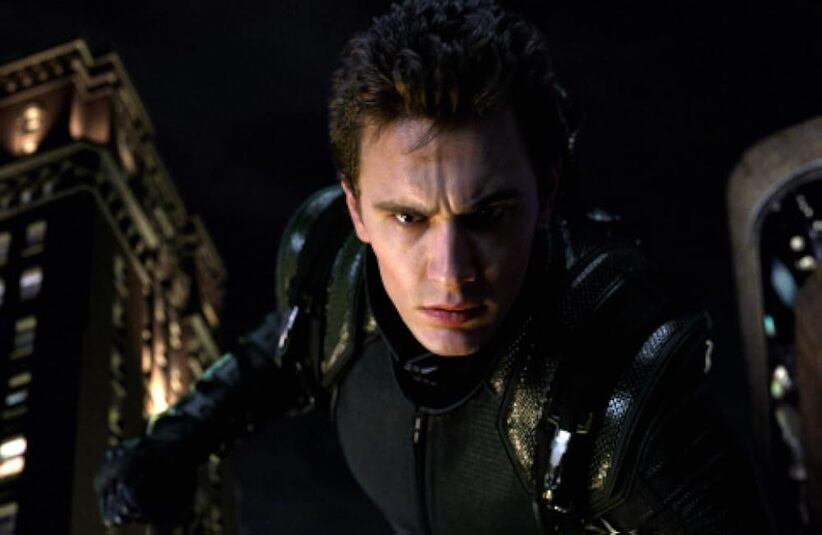 A man in a black armored suit leans forward in a dramatic pose against a dark cityscape. His expression is intense and focused, conveying determination.