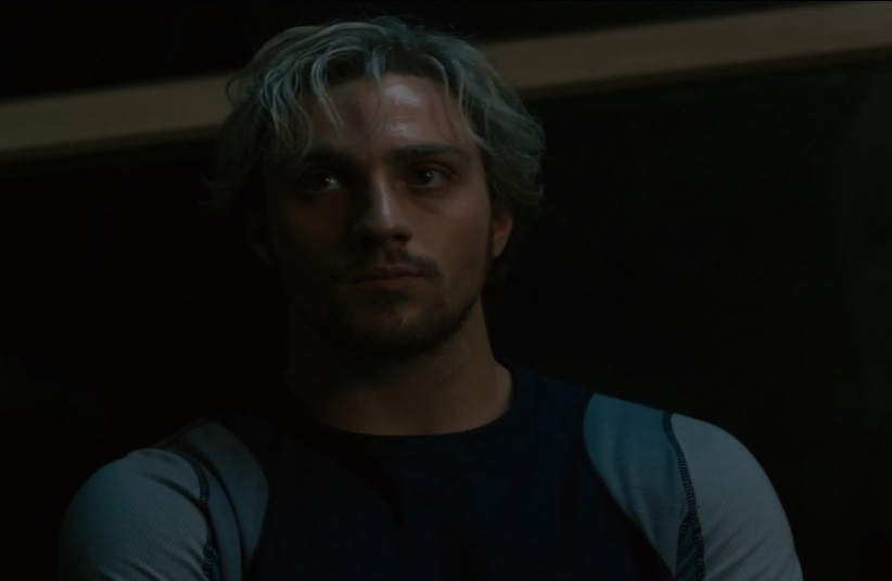 A man with light hair and a serious expression sits in a dimly lit room, wearing a dark shirt with light sleeves, conveying a somber mood.