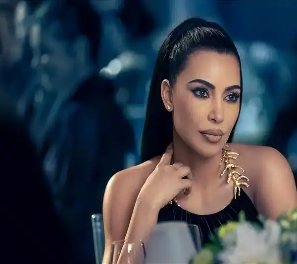 Kim Kardashian in American Horror Story (2011), with a sleek ponytail and black dress, sits thoughtfully at a dimly lit, elegant event. The gold leaf detail on her dress adds a touch of sophistication. PETA