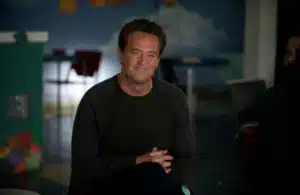 Matthew Perry smiles warmly, sitting indoors with legs crossed. The background includes colorful art and chairs, suggesting a casual setting.