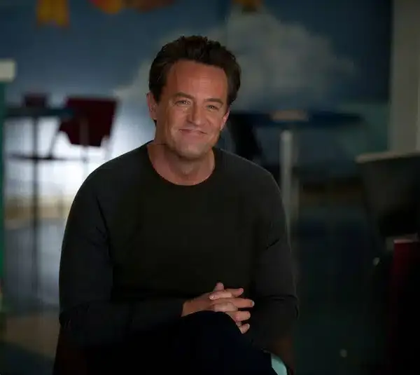 Matthew Perry smiles warmly, sitting indoors with legs crossed. The background includes colorful art and chairs, suggesting a casual setting.
