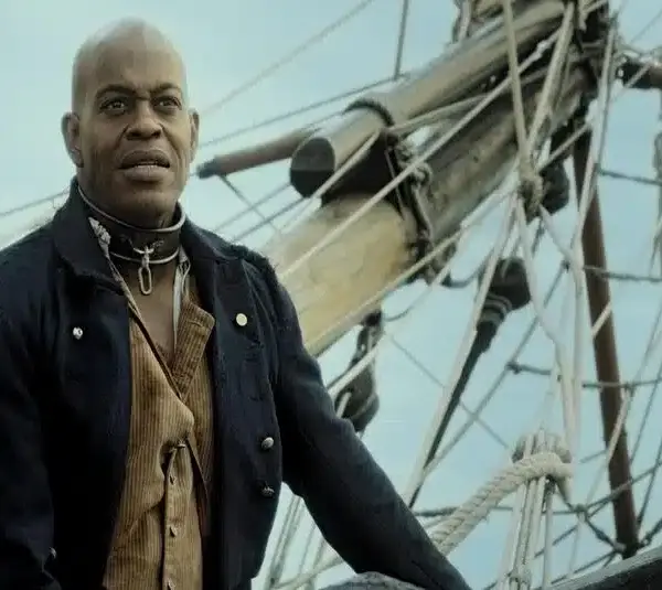 Jubilant Sykes in historical attire stands confidently on a ship's deck, with ropes and wooden masts in the background against a clear sky. The mood is determined.