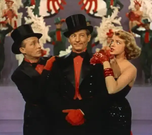 Bing Crosby, Danny Kaye, and Rosemary Clooney in formal black attire and top hats stand on stage, surrounded by dancers in red and green costumes. The scene feels festive and lively.