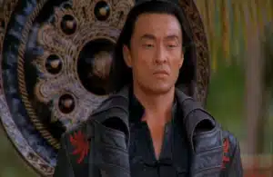Cary-Hiroyuki Tagawa in Mortal Kombat (1995) with long dark hair in a black, ornate coat stands in front of a large decorative shield. He appears serious and calm, surrounded by greenery.