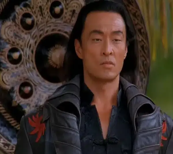Cary-Hiroyuki Tagawa in Mortal Kombat (1995) with long dark hair in a black, ornate coat stands in front of a large decorative shield. He appears serious and calm, surrounded by greenery.