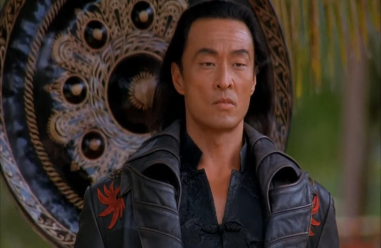 Cary-Hiroyuki Tagawa in Mortal Kombat (1995) with long dark hair in a black, ornate coat stands in front of a large decorative shield. He appears serious and calm, surrounded by greenery.
