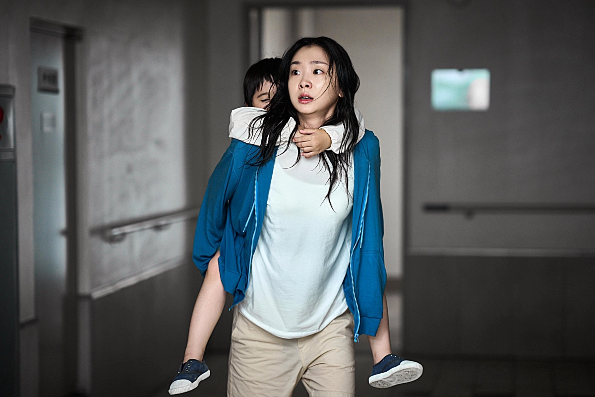 Woman in a blue jacket carries a child on her back in a dim hallway. Her expression is tense and focused, conveying urgency and concern.