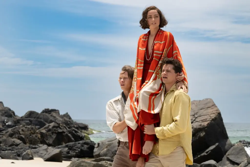Three people stand on a rocky beach. Two men in light shirts lift a woman in a red and orange robe. The sky is clear, suggesting a peaceful day.