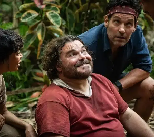 Four people in a jungle setting, one sitting and smiling with a thoughtful expression, others crouched, looking engaged and curious. Lush greenery surrounds them. From the movie Anaconda (2025)