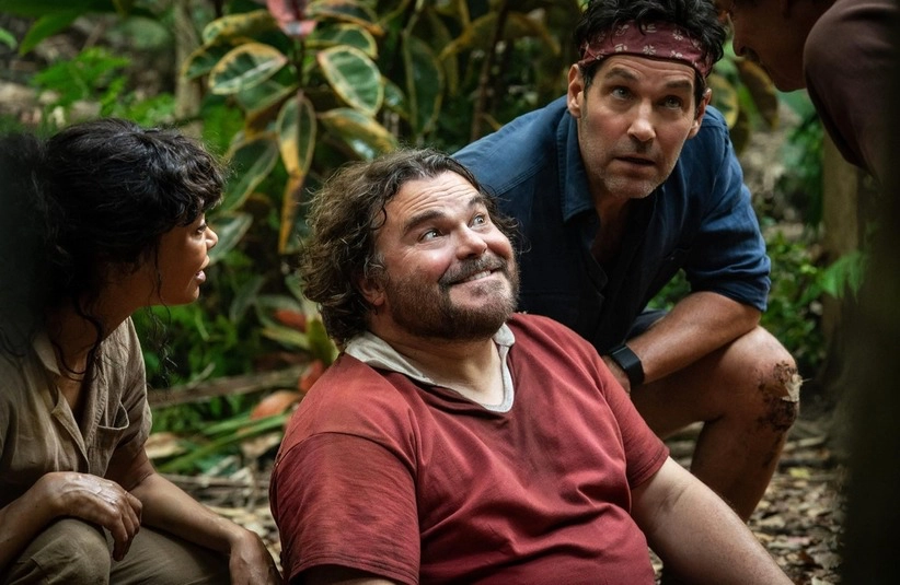 Four people in a jungle setting, one sitting and smiling with a thoughtful expression, others crouched, looking engaged and curious. Lush greenery surrounds them. From the movie Anaconda (2025)