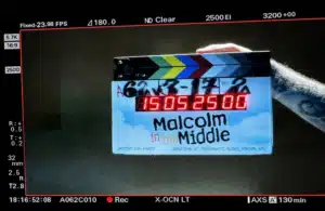 A colorful clapperboard with "Malcolm in the Middle: Life's Still Unfair (2026)" is held by a tattooed arm. The digital display shows numbers under studio lighting.