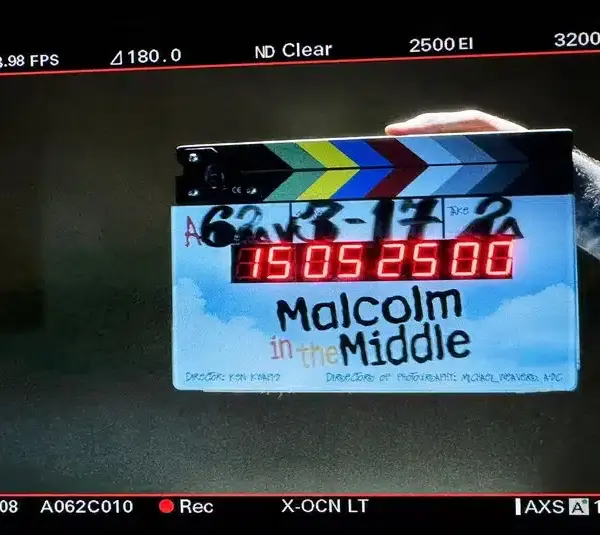 A colorful clapperboard with "Malcolm in the Middle: Life's Still Unfair (2026)" is held by a tattooed arm. The digital display shows numbers under studio lighting.