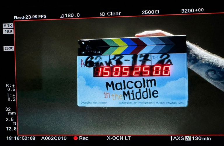 A colorful clapperboard with "Malcolm in the Middle: Life's Still Unfair (2026)" is held by a tattooed arm. The digital display shows numbers under studio lighting.