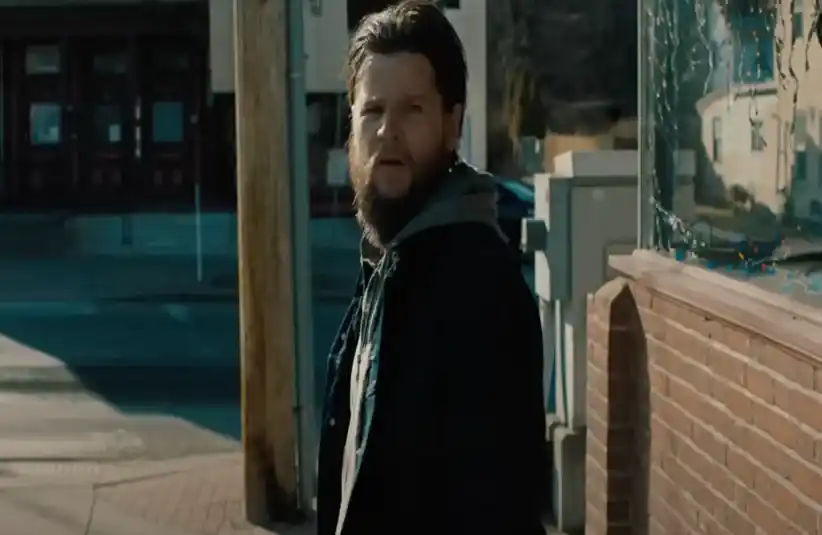 Steven Grayhm in "Sheepdog", as bearded man with long hair stands on a quiet street beside a brick building. He gazes forward intently, creating a tense and contemplative atmosphere.